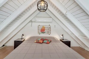 Ein Schlafzimmer im Dachgeschoss mit schrägen Wänden, einem Bett mit einer Steppdecke, zwei Nachttischen mit Lampen, einer Wand-Klimaanlage und einem Wanddekor.