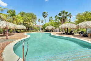 A swimming pool with a curved edge, surrounded by lounge chairs and thatched umbrellas, with palm trees in the background.
