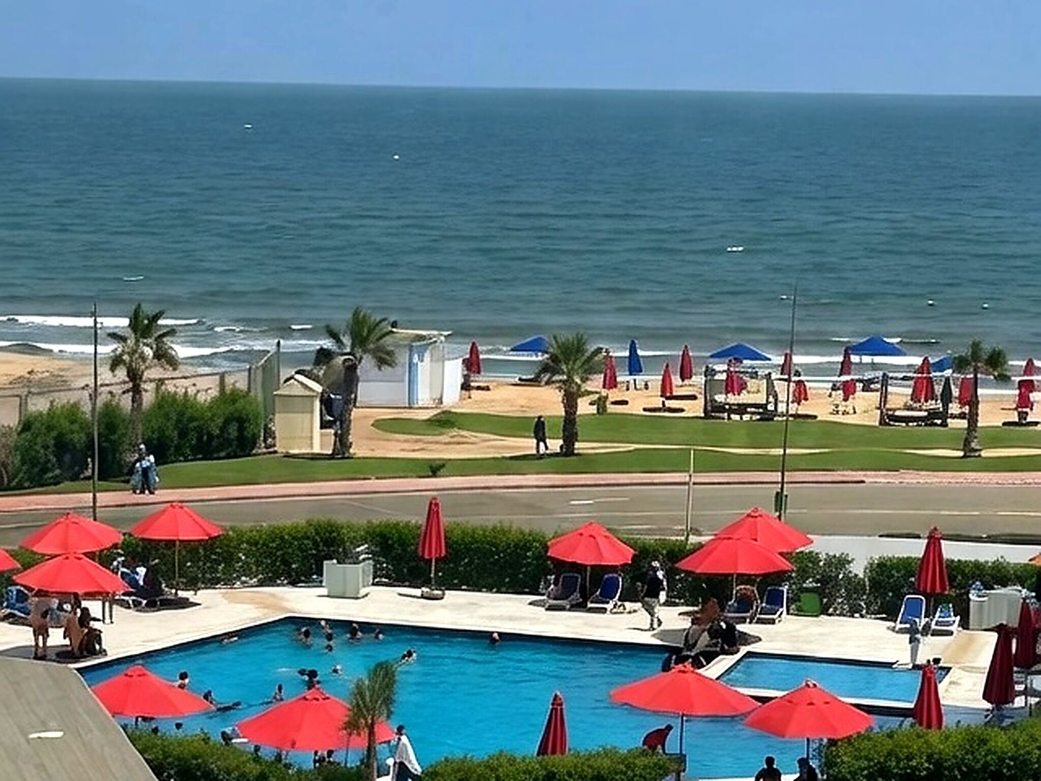 A pool area with red umbrellas, a view of the ocean, and a beach in the background.