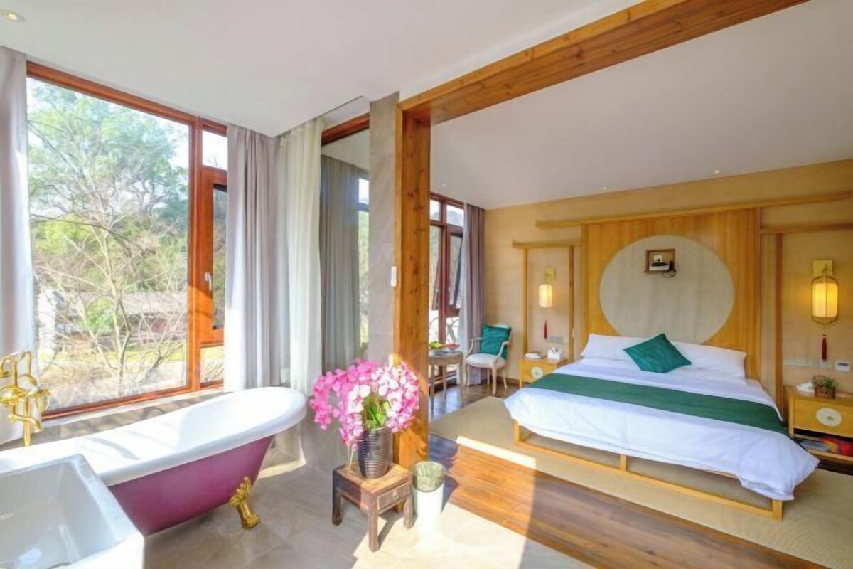 Une chambre avec une grande fenêtre, une baignoire, un lit avec un oreiller vert et une petite table sur laquelle est posé un vase de fleurs.