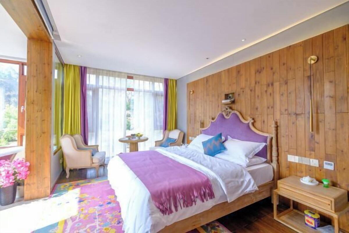Une chambre à coucher avec une tête de lit en bois, un lit recouvert d’une literie blanche et violette, une petite table de chevet et une chaise près de la fenêtre.