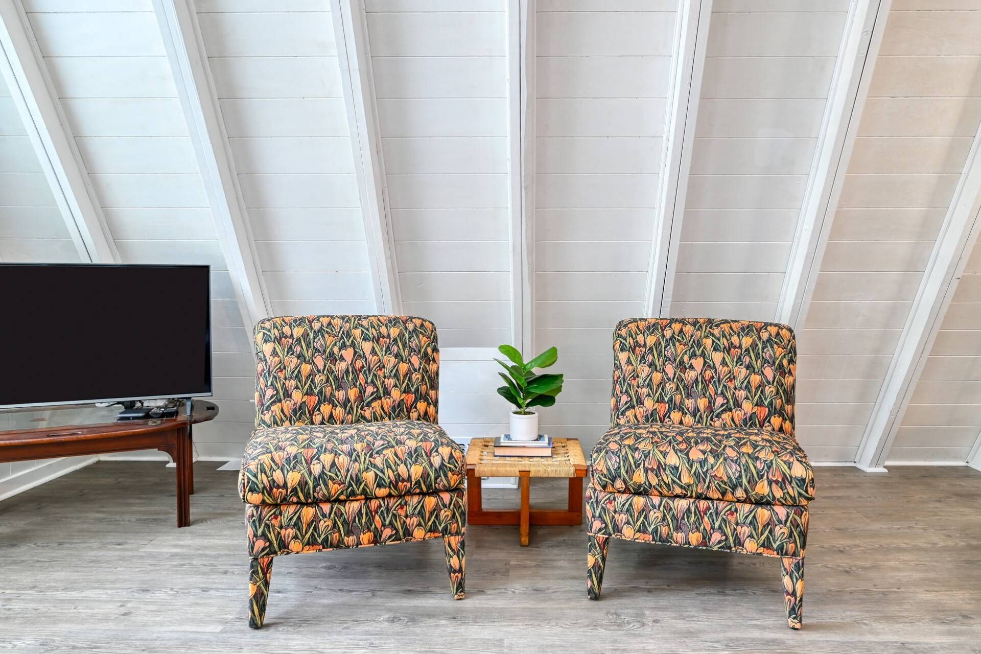 Dos sillones con estampado floral, una mesa de centro de madera, una planta en maceta y un televisor sobre un mueble de madera en una habitación con paredes blancas empapeladas y techo inclinado.