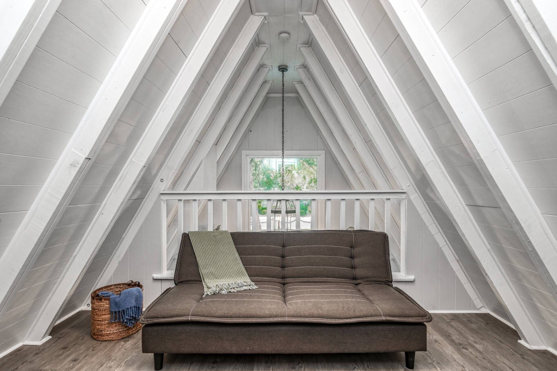 A cozy attic room with a brown sofa, a woven basket, and a view of greenery through a window.
