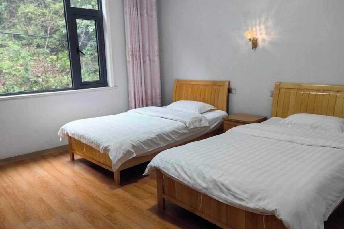 A room with two wooden beds, a window with a view of greenery, and a wall-mounted light fixture.