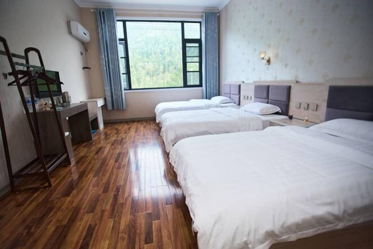 A hotel room with two beds, a wooden floor, a desk with a chair, a window with curtains, and a wall-mounted air conditioner.