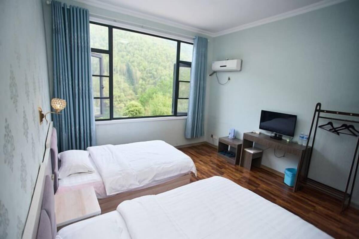 A room with two beds, a TV, a desk, a window with a view of greenery, and a wall-mounted air conditioner.