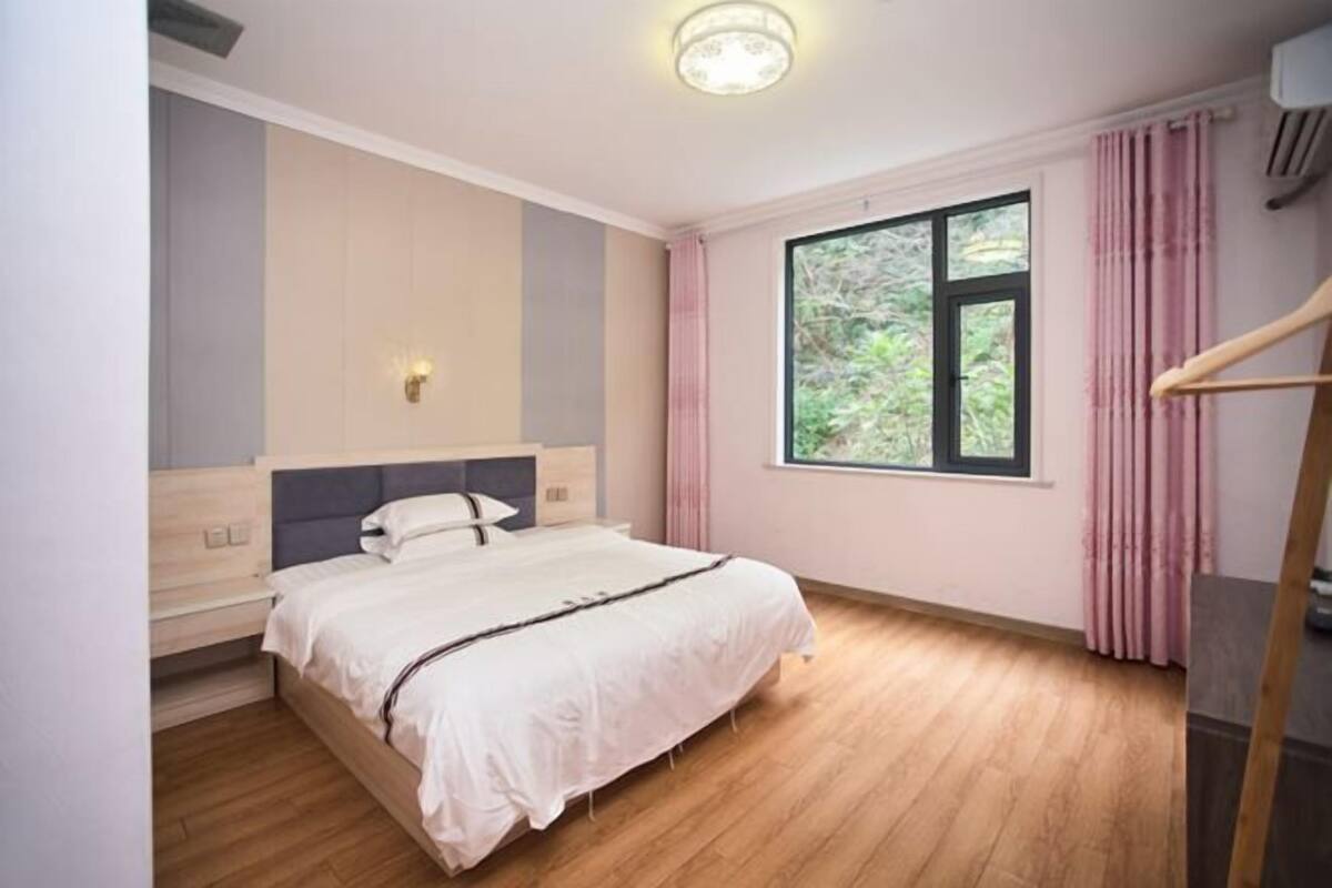 A bedroom with a bed, a window, a wall-mounted light, and a wooden floor.