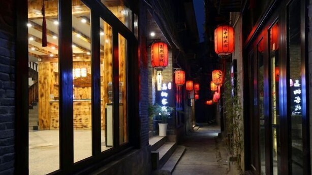 Une ruelle étroite bordée de bâtiments traditionnels en bois, illuminée par des lanternes rouges et une lumière chaleureuse provenant de l’intérieur.