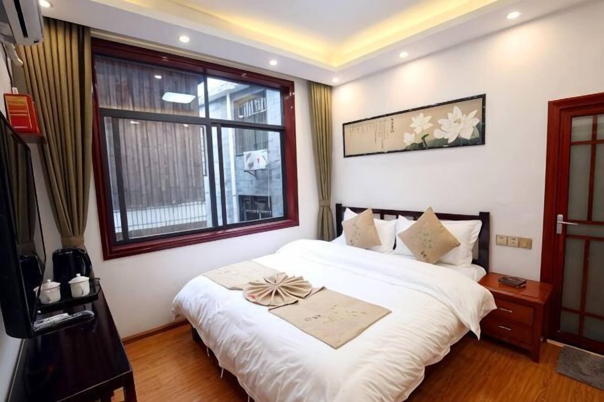 A hotel room with a large bed, a bedside table, a window with curtains, a wall-mounted artwork, and a door.