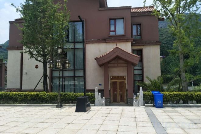 A hotel entrance with a tiled pavement, a wooden door, and a small garden area with a tree and a trash bin.
