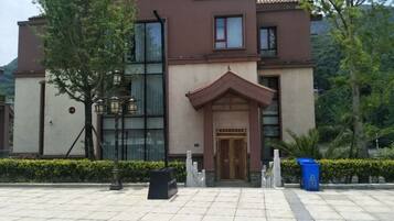A hotel entrance with a tiled pavement, a wooden door, and a small garden area with a tree and a trash bin.