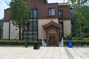 Entrada de hotel con pavimento de baldosas, puerta de madera y un pequeño jardín con un árbol y un contenedor de basura.