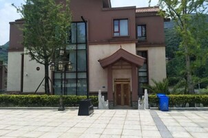 Entrada de hotel con pavimento de baldosas, puerta de madera y un pequeño jardín con un árbol y un contenedor de basura.