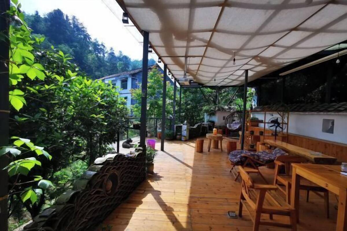An outdoor patio area with wooden benches and tables, surrounded by lush greenery and a stone wall.