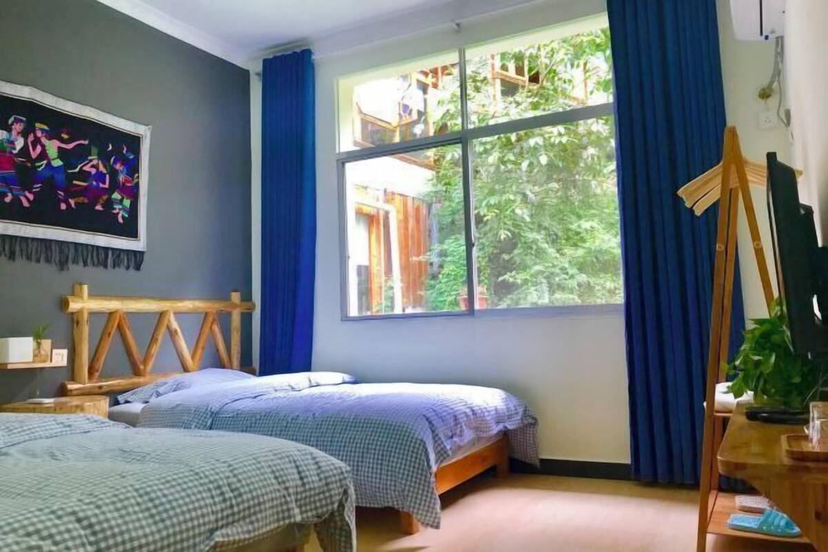 A room with two beds, a large window with blue curtains, a wooden headboard, a painting on the wall, and a desk with a plant.