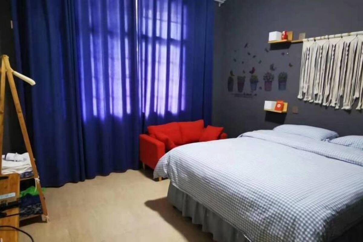 A hotel room with a bed, a red chair, a wooden easel, and a window with blue curtains.