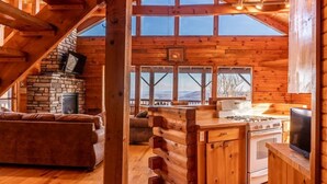 A log cabin interior with a stone fireplace, wooden dining table, and a cozy seating area. - Compton Mountain Cabin w/ Big Mountain Views (Ponca)