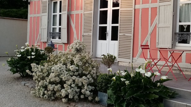 Un bâtiment rose orné de bordures et de volets blancs, un petit jardin avec des fleurs blanches, et une table et une chaise rouges à l’extérieur.