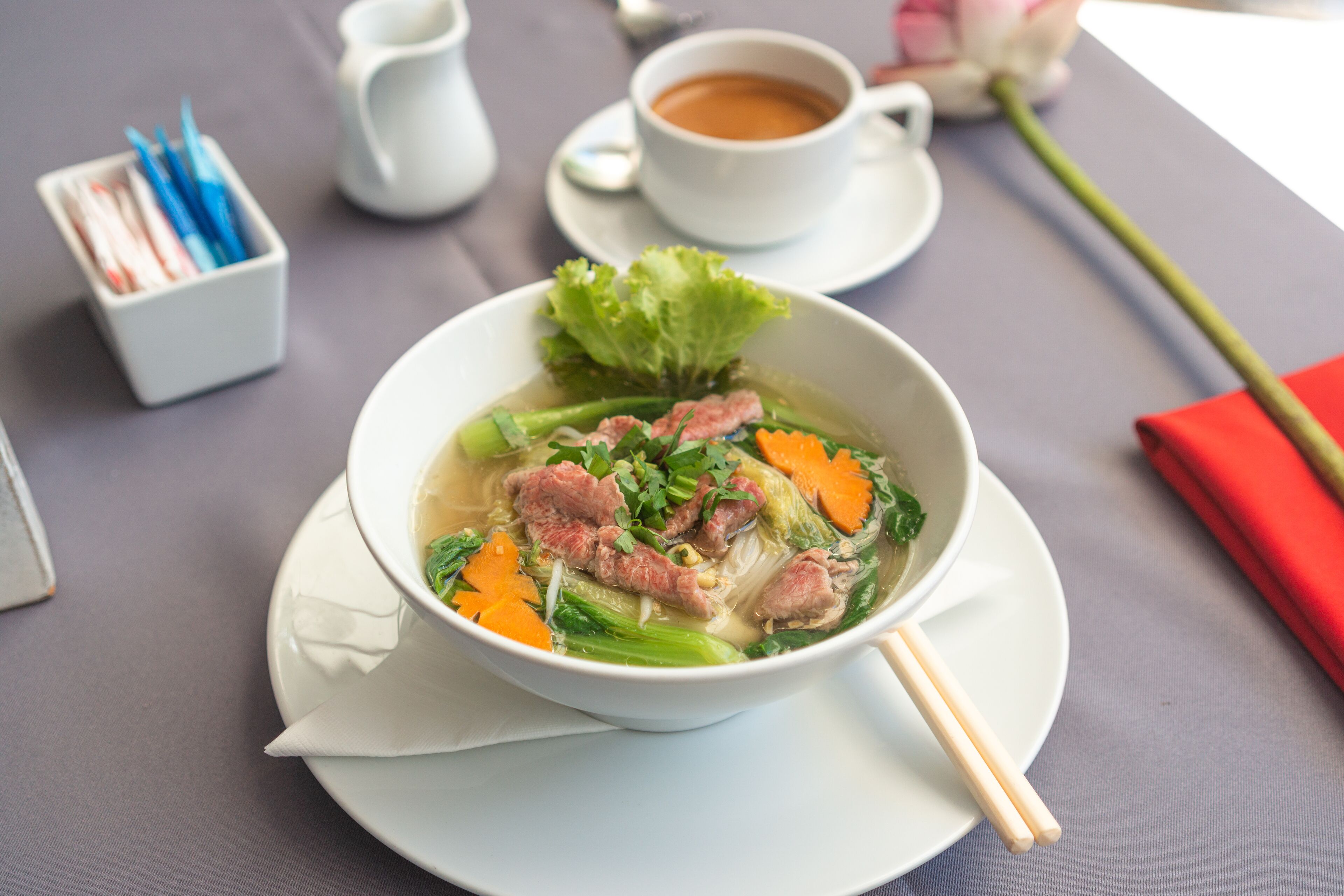 A bowl of soup with vegetables and meat, a cup of tea, chopsticks, and a napkin on a table.