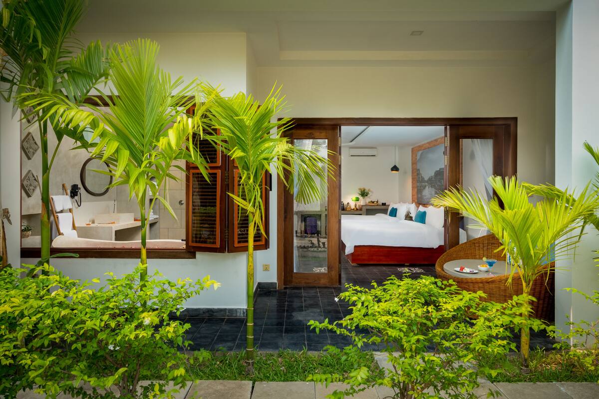 Quarto de hotel com cama, uma pequena mesa de jantar e uma cadeira de vime. Há uma janela grande com persianas de madeira e vista para o jardim.
