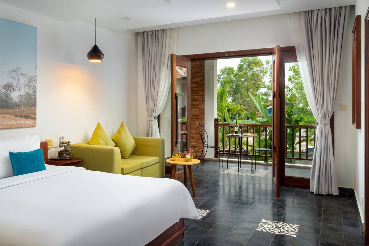 Quarto de hotel com cama, sofá, mesa pequena e varanda com vista para o verde.