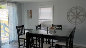 Un comedor con una mesa de vidrio, seis sillas, una lámpara de techo, una ventana con persianas y un adorno de brújula colgado en la pared.