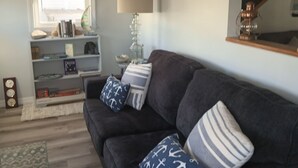 Un salón con un sofá gris oscuro, cojines estampados, una estantería con libros y objetos decorativos, una lámpara y una ventana con persianas.