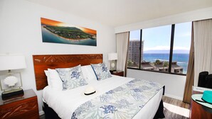 A hotel room with a large bed, a wooden headboard, a painting of a coastal landscape, and a view of a tall building and the sea.