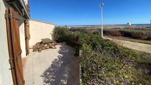 Un patio con una mesa y bancos de madera, una pared blanca con una puerta, y vistas a la playa y las dunas.