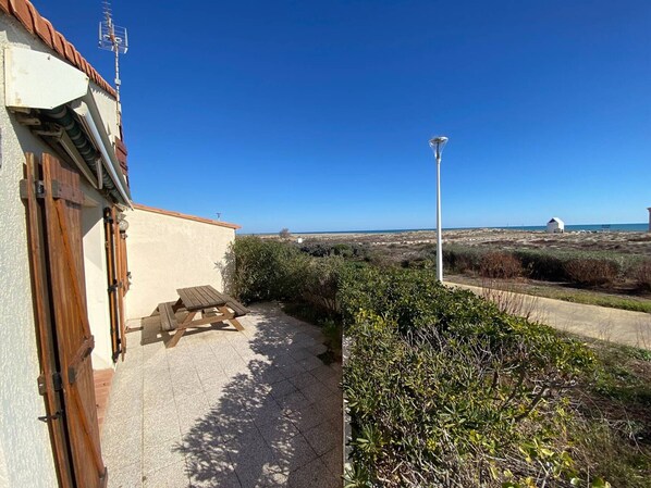 Une terrasse avec une table et des bancs en bois, un mur blanc avec une porte, et une vue sur la plage et les dunes.