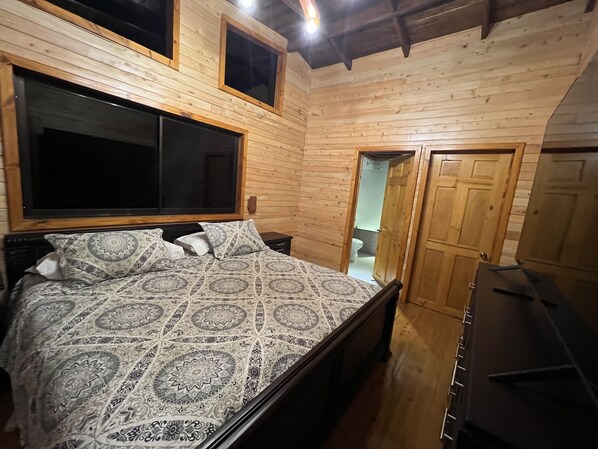A wooden-paneled bedroom with a large bed, two windows, and a door leading to a bathroom.