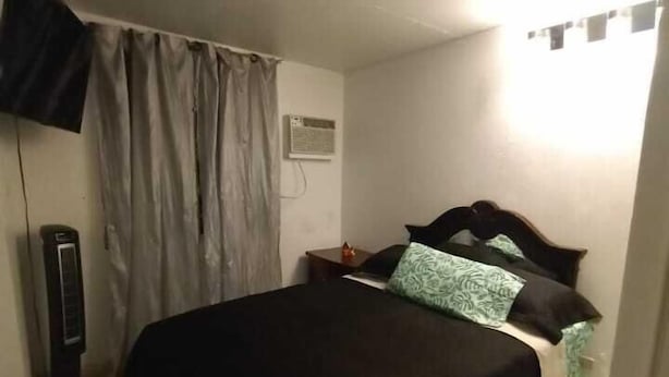 A bedroom with a bed, curtains, a wall-mounted air conditioner, a bedside table, and a TV on the wall. - Cottage at West 7 Palms  (St. Thomas)