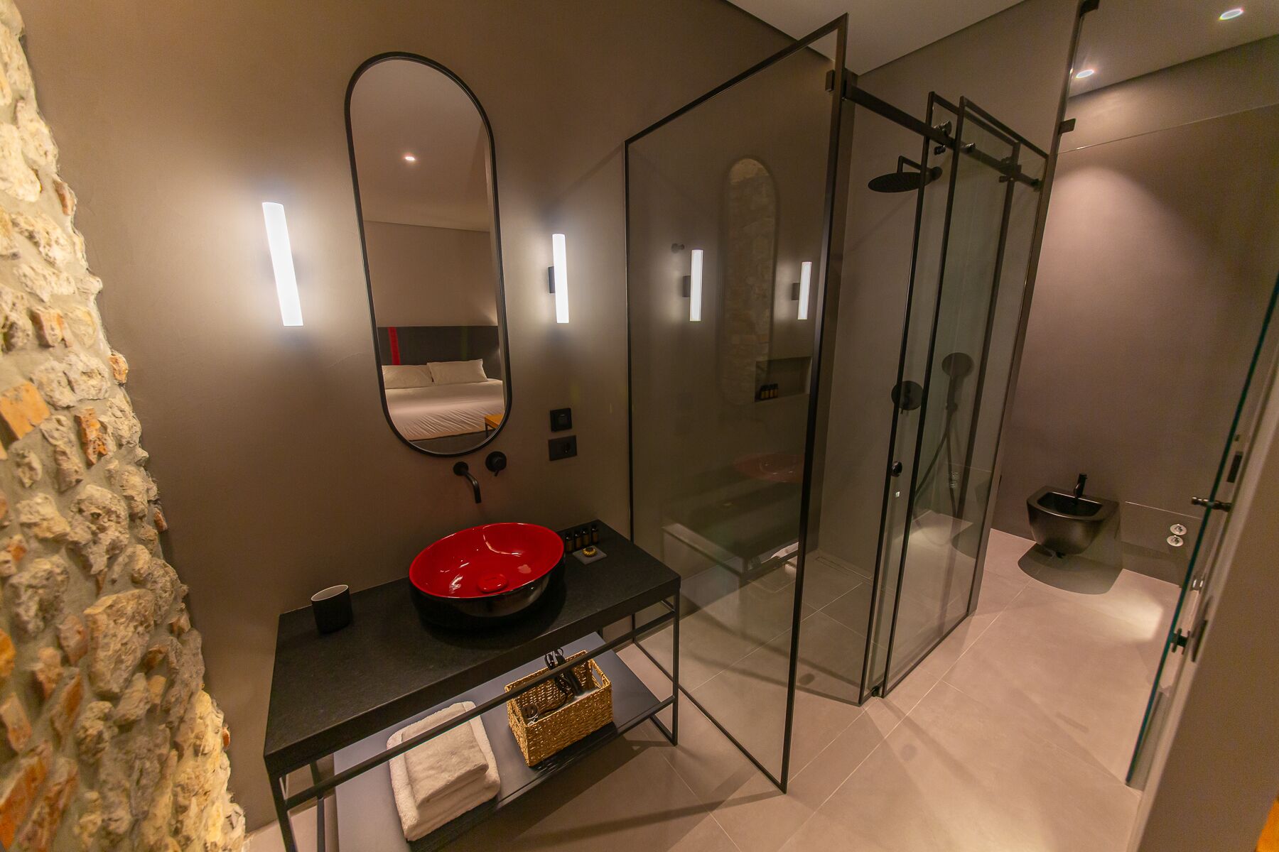 Un baño moderno con ducha envuelta en vidrio, un espejo grande y un lavabo rojo sobre una encimera negra.