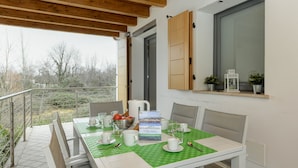 Un coin repas avec une table dressée pour quatre personnes, une fenêtre avec des volets en bois et, en arrière-plan, une vue sur des arbres et des bâtiments.