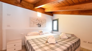 Une chambre à coucher avec une couvre-lit à carreaux, une table de chevet avec une lampe, un tableau de fleurs accroché au mur et une fenêtre donnant sur l’extérieur.