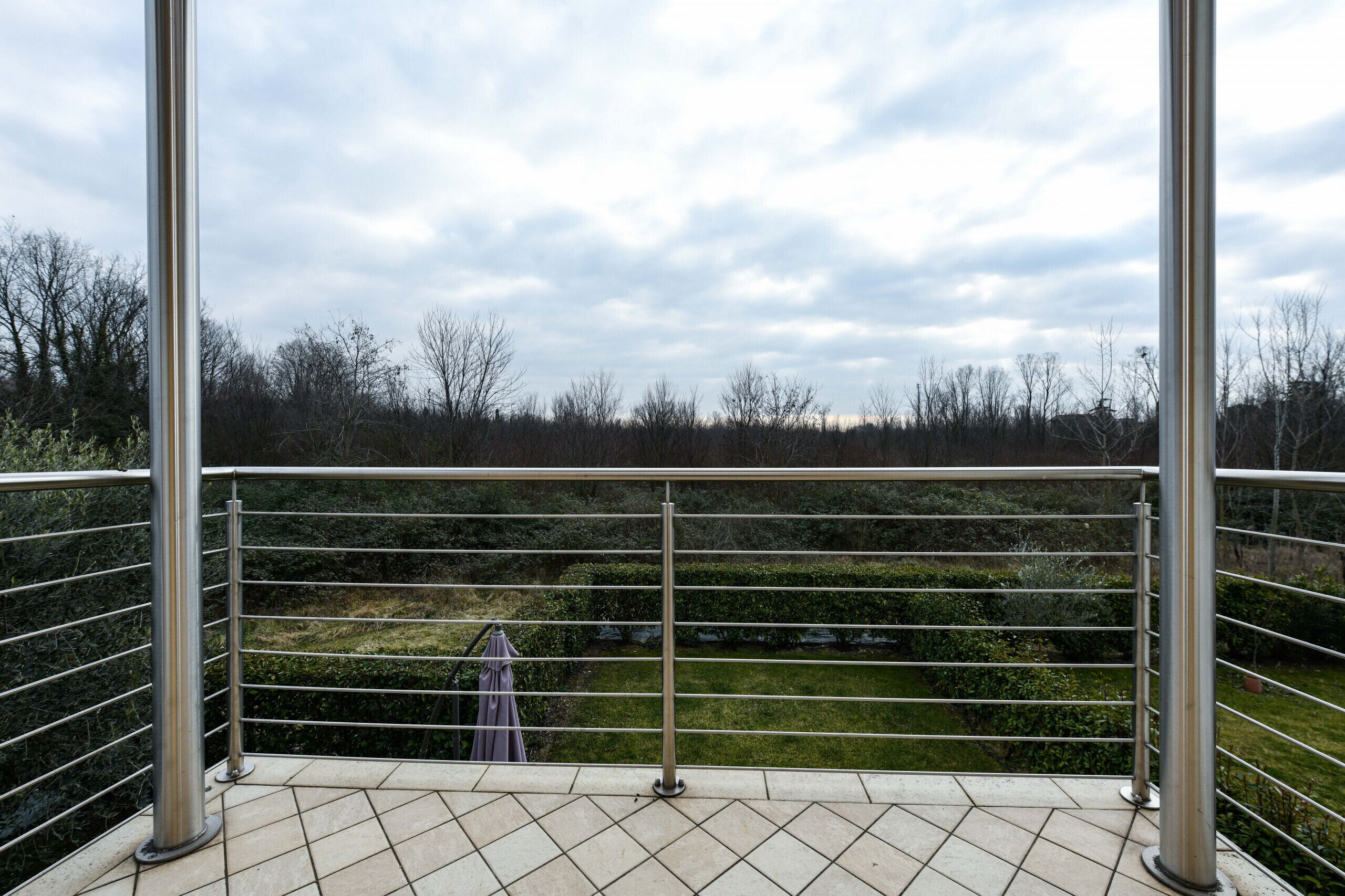 A balcony with metal railings, a tiled floor, and a view of a landscaped garden with trees and bushes.