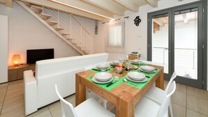 Un coin repas avec une table en bois dressée pour quatre personnes, des chaises blanches, un canapé, un téléviseur et un escalier menant à un niveau supérieur.