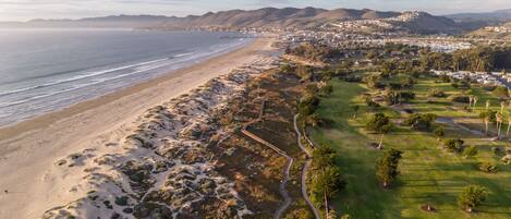 Vista aérea de una zona costera con playa, campo de golf y área residencial con colinas al fondo.