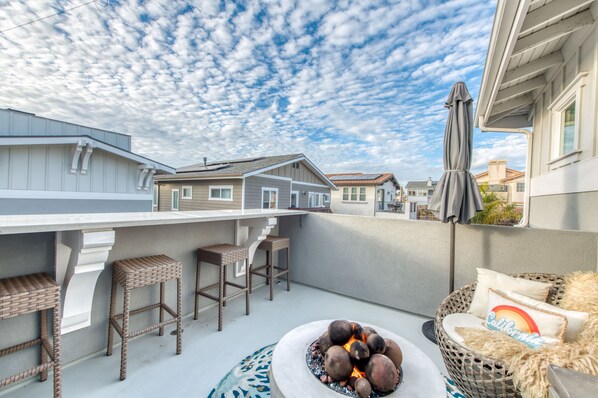 A patio with a fire pit, wicker bar stools, and a wicker chair with a cushion.