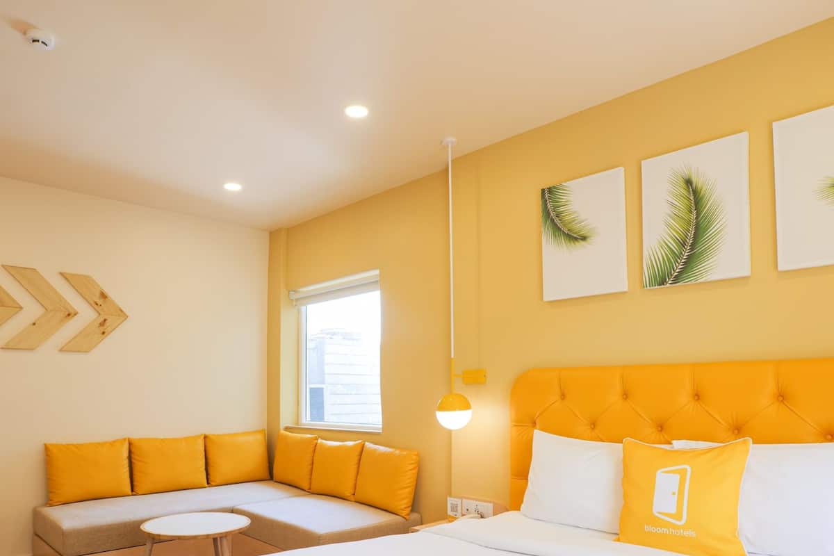 Ein Hotelzimmer mit einem Bett, einem Sofa mit gelben Kissen, einem kleinen Tisch und einem Fenster mit Jalousien.