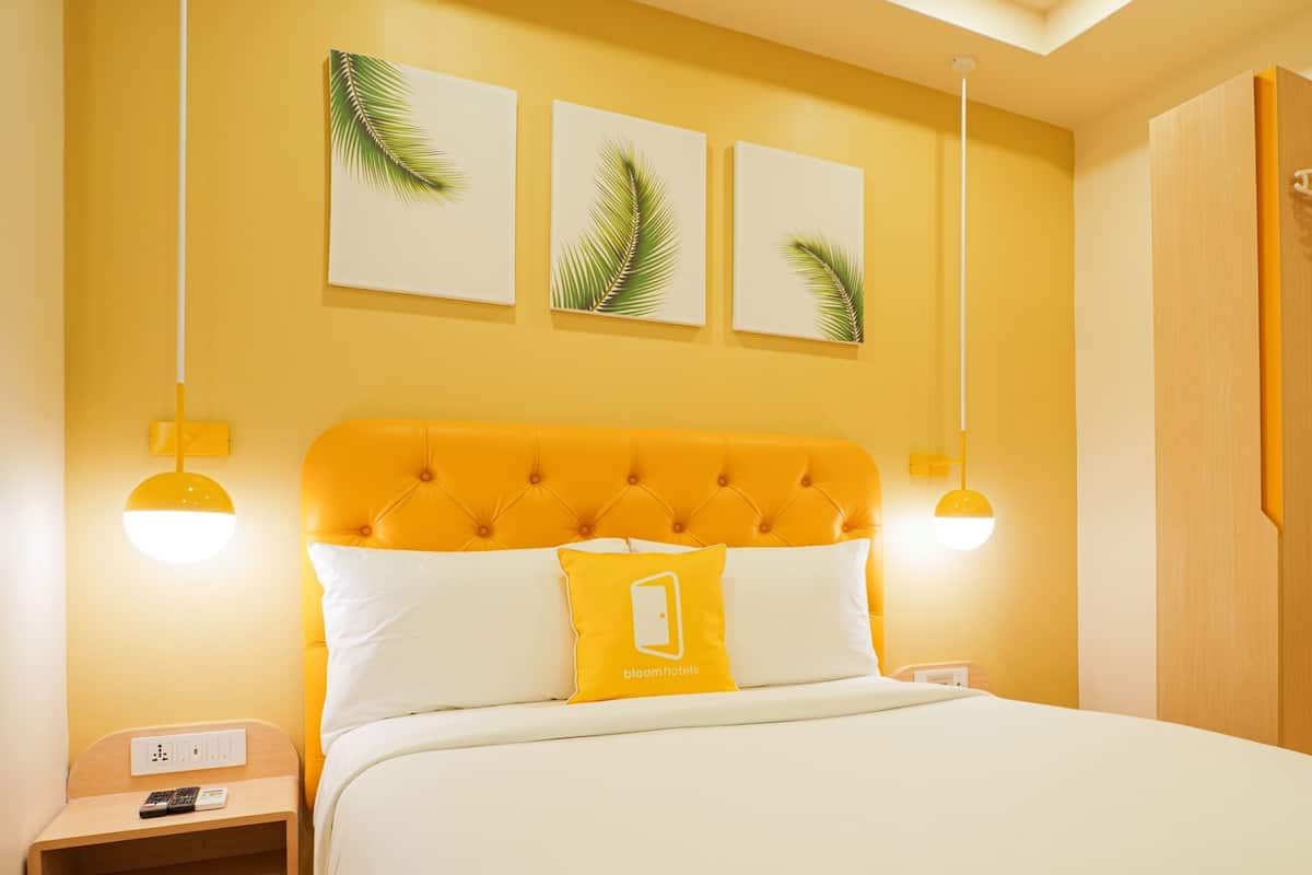 Ein Hotelzimmer mit gelber Wand, einem gesteppten Kopfteil, einem Bett mit weißer und gelber Bettwäsche sowie drei Wandbildern aus Palmblättern.
