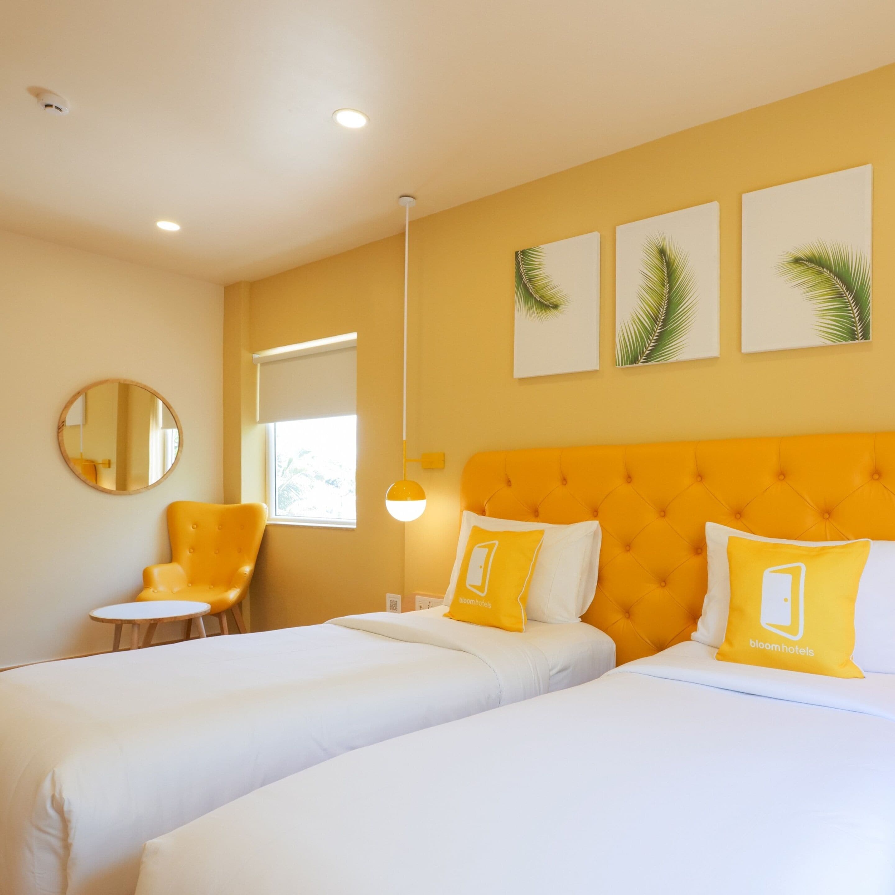 A hotel room with two beds, a round mirror, a chair, and a window with blinds.