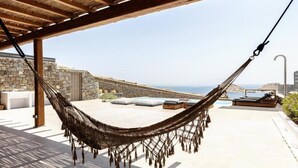 Una hamaca bajo una pérgola de madera, con vistas a una zona de piscina con sillones de descanso y un muro de piedra.