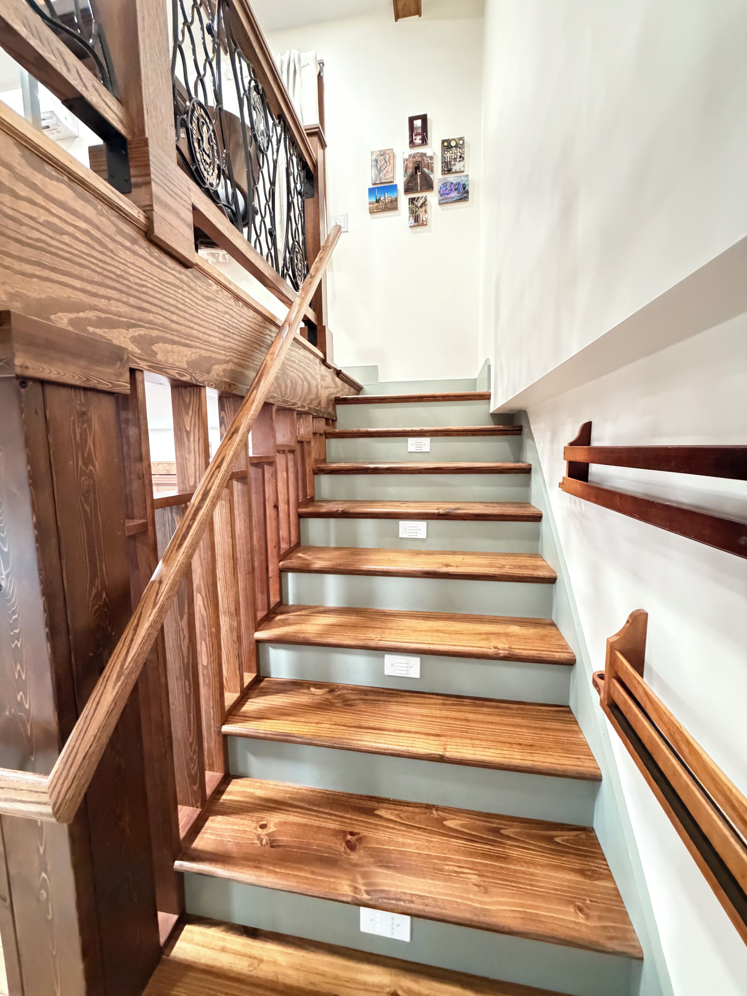 A wooden staircase with a metal railing, leading to a landing with a white wall and several framed pictures.