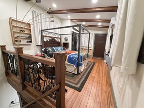 A bedroom with a four-poster bed, wooden flooring, a small bedside table, and a shelf with decorative items.