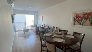 Ein Essbereich mit einem runden Tisch, gedeckt für vier Personen, umgeben von Stühlen. Im Hintergrund befindet sich eine Couch und ein kleiner Tisch, an der Wand hängt ein Gemälde von Tulpen.