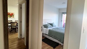 Ein Schlafzimmer mit Bett, Nachttisch und Fenster. Ein Flur führt zu einem Essbereich mit Tisch und Stühlen.