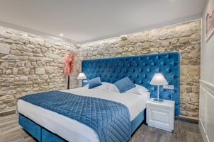 A bedroom with a stone wall, a blue upholstered headboard, a quilted bedspread, two bedside lamps, and a nightstand.