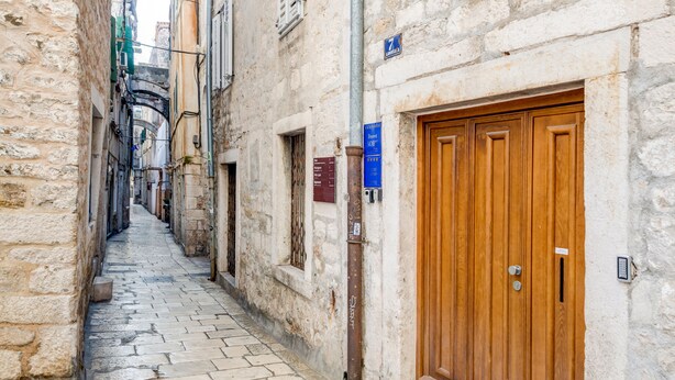 A narrow stone-paved alleyway with a wooden door on the right, flanked by buildings with visible signs and a metal pole. - Mint Luxury Rooms (Split)