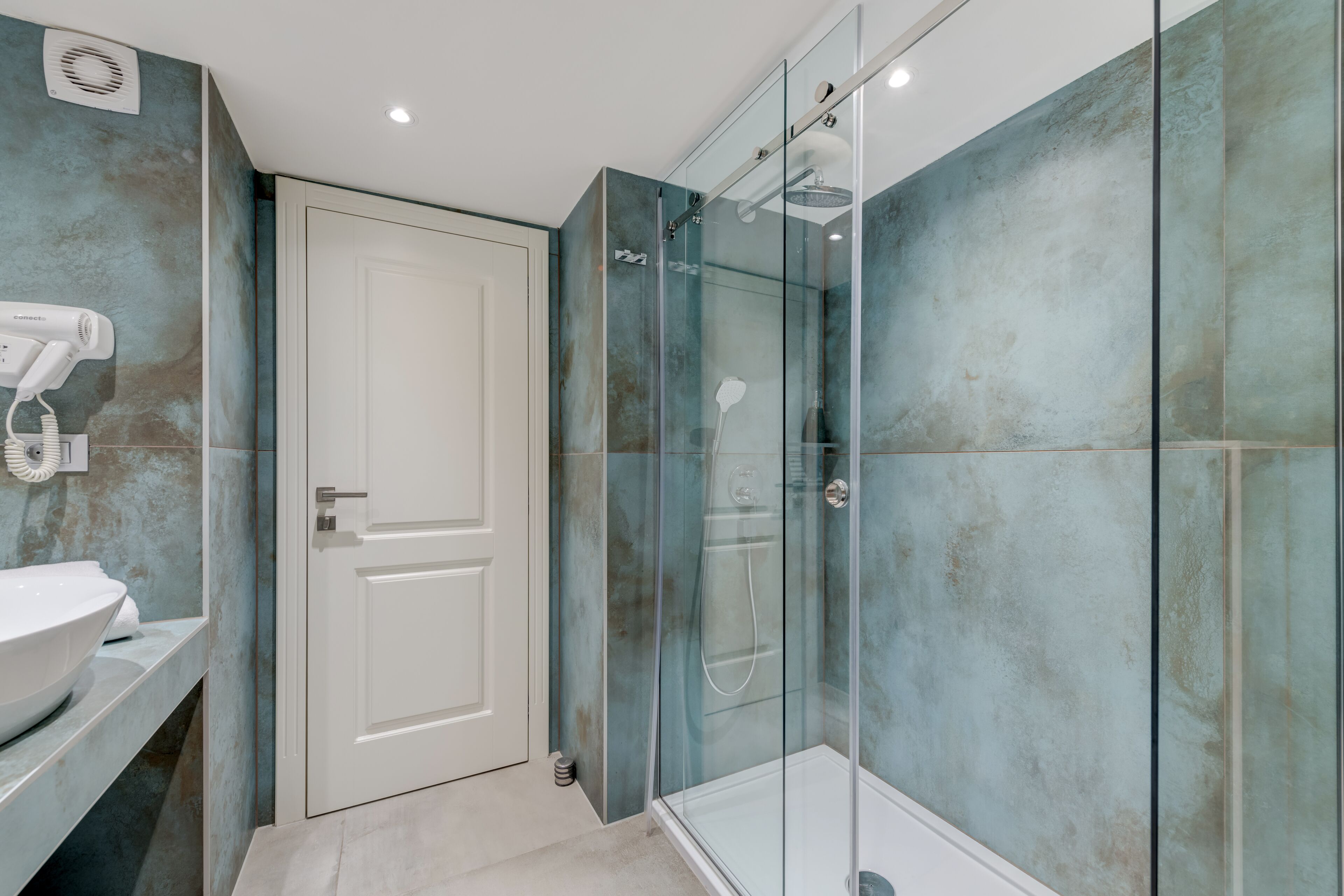 Une salle de bain avec une douche à parois de verre, un lavabo blanc et un sèche-cheveux fixé au mur.